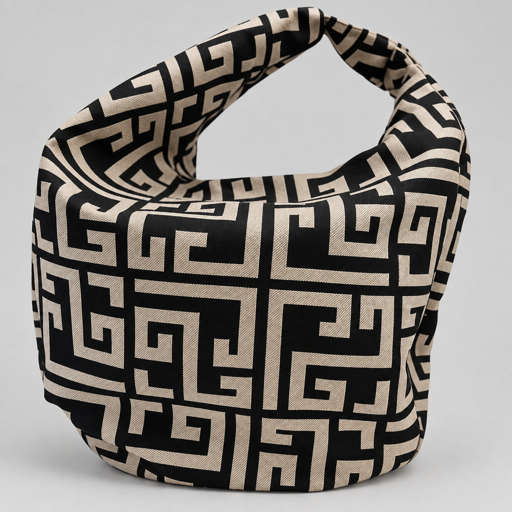 A black and beige geometric patterned shoulder bag with a loop handle, displayed on a white surface with a blurred boutique background.