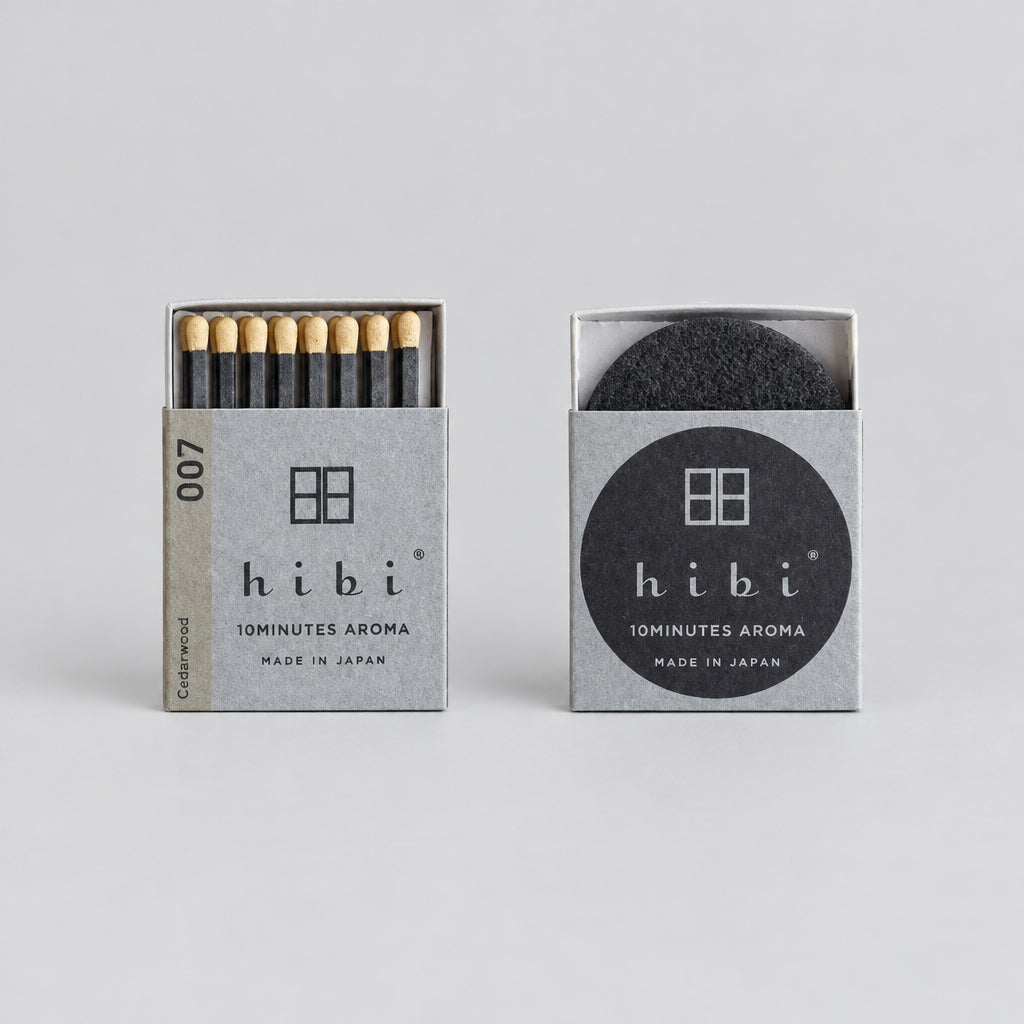 Two Hibi 10Minutes Aroma matches on a wooden surface, showing both the silver and black outer packaging.