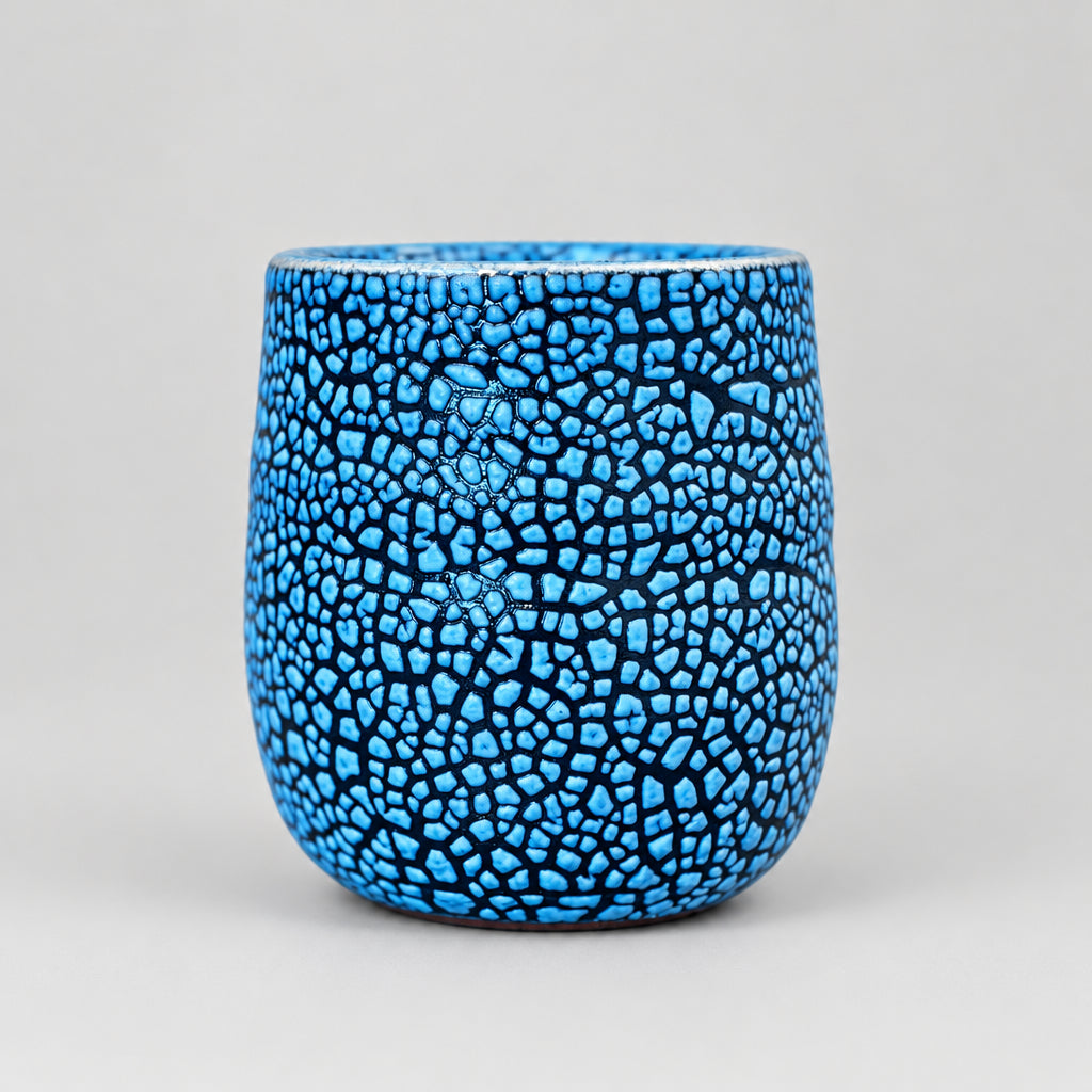 A small ceramic cup with a vibrant blue and black crackle glaze pattern, sitting on a textured gray stone surface with a blurred outdoor background.