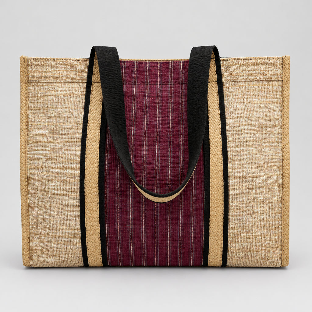 A woven tote bag made from natural jute with a central burgundy and white striped fabric panel and black handles, resting on a white surface with a blurred indoor background.